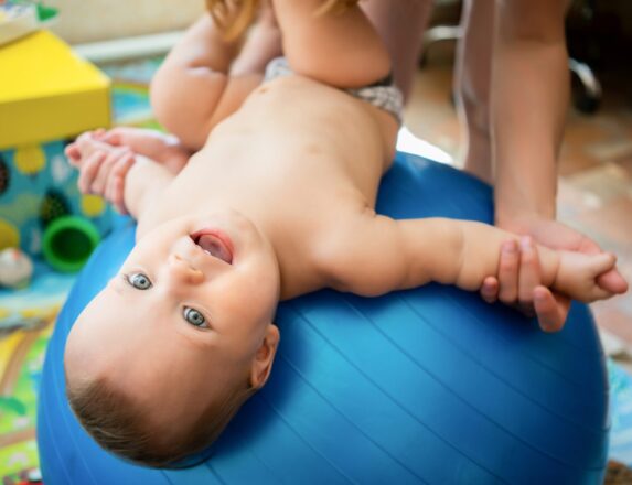 Top view of little caucasian kid enjoying exercises on big blue fitness ball with unrecognizable mother, looking at camera and smiling. Woman training attractive naked baby in nursery.